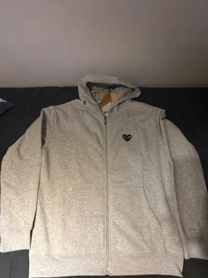 Grå hoodie med dragkedja och hjärtdetalj - Säljer en stilren grå hoodie med dragkedja och huva. På bröstet finns en svart hjärtdetalj med ögon, vilket ger en unik touch. Hoodien har fickor framtill och ribbade muddar vid ärmar och nederkant. Perfekt för en avslappnad look.