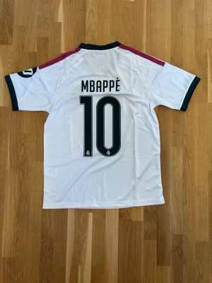 Real Madrid Mbappé matchtröja vit - Snygg vit Real Madrid matchtröja med Mbappé och nummer 10 på ryggen. Tröjan har svarta detaljer vid krage och ärmslut, samt vinröda ränder på axlarna. Adidas-logga och Real Madrid-emblem på bröstet, samt Emirates Fly Better-tryck framtill.