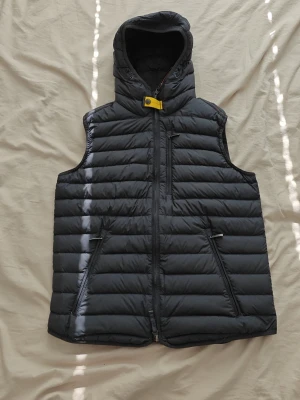 Parajumpers väst med huva - Snygg svart dunväst med huva och gul detalj vid halsen. Västen har dragkedja framtill, två snedställda fickor med dragkedja och en bröstficka. Insidan är ljusgrå med orange detaljer och etiketter. Perfekt för lager-på-lager och håller dig varm.