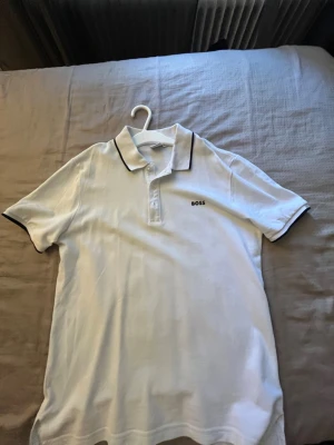 Svart pikétröja från BOSS 220kr styck!! - Klassisk svart pikétröja från BOSS med vit logga på bröstet och vita detaljer längs krage och ärmslut. Tröjan har korta ärmar, knappar vid halsen och är tillverkad i ett mjukt material som känns bekvämt mot huden. 220kr styck!!
