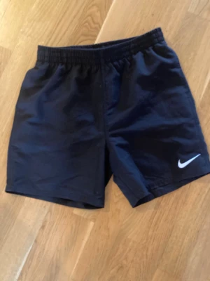 Svarta sportiga shorts från Nike - Säljer ett par svarta sportiga shorts från Nike med elastisk midja och den klassiska vita swoosh-loggan på benet. Perfekta för träning eller chill dagar. Materialet känns lätt och bekvämt.