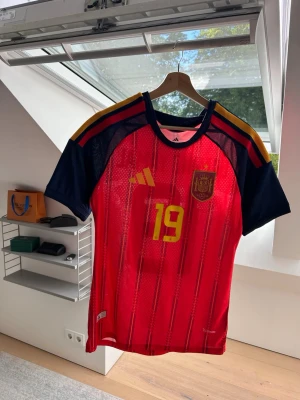 Spansk landslagströja Adidas röd - Säljer en röd spansk landslagströja från Adidas med nummer 19 i gult på bröstet. Tröjan har korta ärmar i marinblått med gula och röda ränder på axlarna samt klubbmärke på vänster bröst. Tillverkad i lätt och ventilerande polyester.