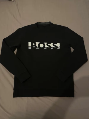 Svart stickad tröja från BOSS - Snygg svart stickad tröja från BOSS med vit logga i grafisk design på bröstet. Tröjan har rund halsringning och långa ärmar. Perfekt för dig som gillar stilrena och moderna plagg.