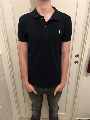 Mörkblå pikétröja från Polo Ralph Lauren - Klassisk mörkblå pikétröja från Polo Ralph Lauren med vit broderad logga på bröstet. Tröjan har korta ärmar, krage och knappar framtill. Perfekt till jeans eller shorts för en clean och stilren look.