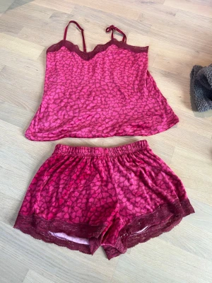 Rosa/rött pyjamasset med spetsdetaljer - Sött pyjamasset i rosa med hjärtmönster från hunke Möller, består av ett linne med tunna axelband och matchande shorts. Båda delarna har mörkare spetsdetaljer längs kanterna.