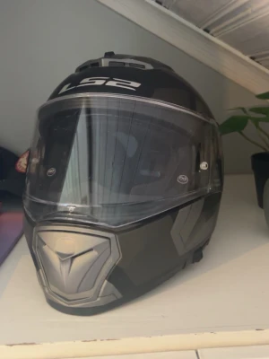 Svart och grå MC-hjälm LS2 - Säljer en svart och grå motorcykelhjälm från LS2 med geometriskt mönster och klart visir. Hjälmen har en modern design och tydliga ventilationsöppningar. Perfekt för dig som vill ha både stil och säkerhet på vägen. 