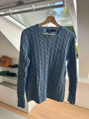 Blå kabelstickad tröja från Polo Ralph Lauren - Säljer en blå kabelstickad tröja från Polo Ralph Lauren. Tröjan har rund halsringning, lång ärm och klassisk kabelstickad design. Den har en liten broderad logga på bröstet och ribbade muddar vid ärmslut och nederkant. Perfekt för dig som gillar stilrena plagg.