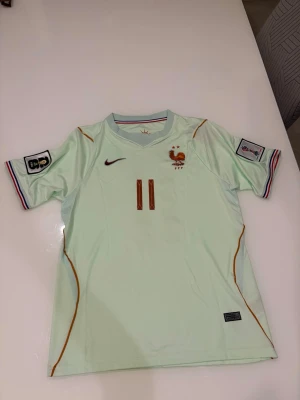 Ljusblå Frankrike fotbollströja - Säljer en mintgrön fotbollströja med nummer 11 och namnet Olise på ryggen. Tröjan har Frankrikes landslagsemblem, Nike-logga och detaljer i guld och rött. Korta ärmar med randiga muddar och patchar på båda ärmarna. Perfekt för match eller träning. Tröjan är i helt nytt skick och kommer i storlek M.