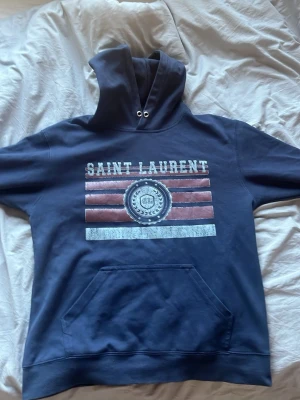 Mörkblå hoodie med tryck - Snygg mörkblå hoodie med stor huva och magficka. Framsidan har ett tryck med texten 'Saint Laurent' och röda samt vita ränder, samt ett emblem i mitten. Hoodien har ribbade muddar och dragsko i huvan. Perfekt för en avslappnad streetwear-look.