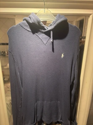 Blå hoodie från Polo Ralph Lauren - Säljer en blå hoodie från Polo Ralph Lauren i ett strukturerat, waffle-mönstrat material. Hoodien har huva med dragsko och den klassiska broderade loggan på bröstet. Perfekt för en avslappnad och stilren look.