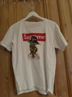 Vit Supreme t-shirt med Yoda-tryck - Säljer en vit t-shirt med Supreme-logga och en cool Yoda-illustration på framsidan. T-shirten har korta ärmar och en klassisk passform. Perfekt för dig som gillar streetwear och vill sticka ut med en unik design.