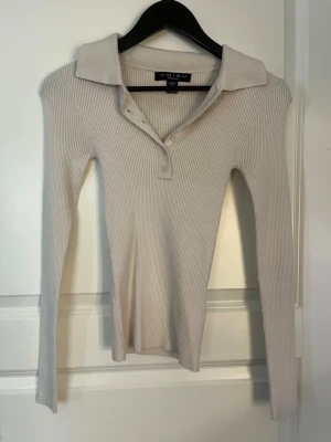 Beige ribbad långärmad polotopp - Snygg beige ribbad polotopp med lång ärm och klassisk krage. Toppen har knappar framtill och är i stretchigt material som sitter snyggt på kroppen. Perfekt att styla med jeans eller kjol. Aldrig anvönd.
