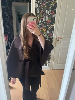 Mörkbrun cape-kappa med skärp - Snygg mörkbrun cape-kappa med bred krage och knytband i midjan. Kappan har vida ärmar och en avslappnad passform som ger en cool siluett. Perfekt för dig som vill ha något annorlunda och trendigt i garderoben. 