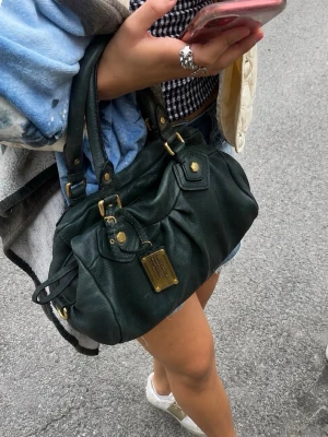 Mörkgrön handväska i skinn - Snygg mörkgrön handväska i skinn från Marc by Marc Jacobs med guldfärgade metalldetaljer och dubbla handtag. Väskan har en dekorativ metallbricka framtill och ett foder med grafiskt svartvitt mönster med märkets logga. Perfekt för dig som gillar trendiga accessoarer.