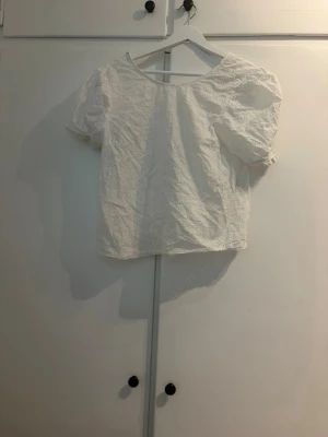 Vit blus med puffärm och broderi - Söt vit blus med puffiga korta ärmar och broderade detaljer. Blusen har en rak passform och är tillverkad i ett lätt bomullstyg som känns luftigt. Perfekt för dig som gillar romantiska och feminina toppar.