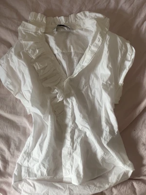 Vit blus med volangdetalj - Stilren vit blus med korta ärmar och volang längs knäppningen. Blusen har en lätt v-ringning och är gjord i ett tunt, mjukt tyg som ger en luftig känsla. Perfekt för en trendig och avslappnad look.