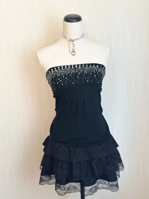 Svart tubtopp med strass från Hello Kiss! - Svart tubtopp från Hello Kiss! med glittriga strassdetaljer upptill. Toppen är axelbandslös och har en figurnära passform, perfekt för en trendig och elegant stil.