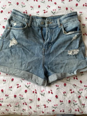 Blå denimshorts - Säljer ett par klassiska blå denimshorts med hög midja och uppvikta benslut.  Storlek 38, uppvikningarna behöver strykas efter att de tvättades & nu inte är perfekt vikta längre.