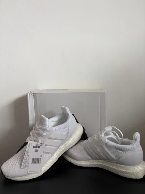 Vita Adidas Ultraboost sneakers - Snygga vita Adidas Ultraboost sneakers med stickad ovandel i mesh för grym komfort och andningsförmåga. Skorna har klassiska tre ränder på sidan, vit Boost-sula och svart yttersula. Perfekta för dig som gillar stilrena och sportiga sneakers.