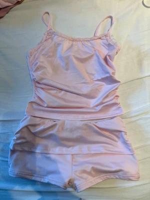 Ljusrosa linne och shorts set - Sött ljusrosa underklädesset med linne och matchande shorts. Linnet har tunna axelband och spetsdetaljer vid urringningen. Materialet är mjukt och glansigt, troligen polyester eller satinliknande tyg. Perfekt för en mysig kväll hemma.