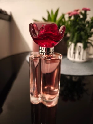 Oscar de la Renta Esprit d’Oscar Amour - En elegant parfymflaska med ljusrosa doft och en unik röd blomformad kork. Flaskan är genomskinlig och har mjuka, feminina former. Parfymen kommer i en matchande rosa förpackning med blommigt motiv. Perfekt för dig som gillar lyxiga och romantiska dofter.