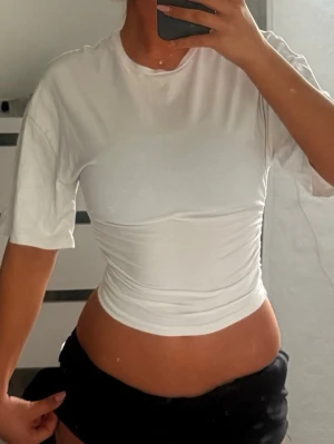 Vit croppad topp med rynkade sidor - Säljer en vit croppad topp med rynkade sidor och korta ärmar. Toppen har en tight passform och är tillverkad i ett mjukt bomullsliknande material. Perfekt till jeans eller kjol för en clean och fräsch look.