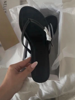 Svarta flip-flop sandaler - Stilrena svarta flip-flop sandaler med tunna remmar och vit kontrastsöm. Klassisk design med platt sula, perfekta för sommaren. Materialet ser ut att vara syntetiskt och remmarna har en diskret detalj i vitt.