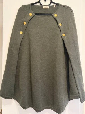 Stenströms Mary mörkgrön merinoull cape - Stilren mörkgrön cape med sex dekorativa guldknappar framtill. Capen har en vid och svepande passform och är tillverkad i merinoull. Perfekt för lager på lager och ger en elegant touch till din outfit.
