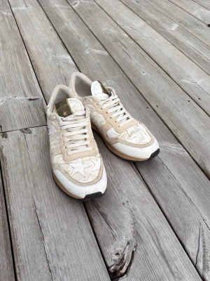 Rockrunner Valentino Garavani sneakers med stjärnor - Säljer ett par snygga Rockrunner Valentino Garavani sneakers i beige och vitt med stjärnmönster på sidorna. Skorna har detaljer i mocka och läder, vita snören och en cool nitar-detalj på hälen. Perfekta för dig som gillar exklusiva och unika sneakers.