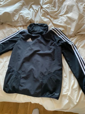 Svart träningsjacka från Adidas - Svart träningsjacka från Adidas med vita ränder längs ärmarna och logga på bröstet. Jackan har hög krage, tumhål i ärmsluten och är tillverkad i funktionsmaterial. Perfekt för träning eller sportaktiviteter.