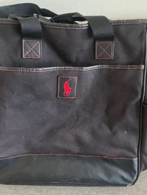 Svart axelremsväska med röd ralph lauren logga - Snygg svart axelremsväska från Polo Ralph Lauren med röd broderad logga framtill. Väskan har både handtag och axelrem, samt röda kontrastsömmar. Tillverkad i slitstark canvas med detaljer i konstläder. Perfekt för dig som vill ha en stilren och rymlig väska. ca 38x35cm 