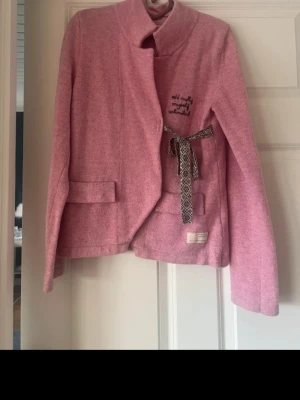 Rosa kofta från Odd Molly - Säljer en rosa kofta från Odd Molly med broderad text på bröstet och ett snyggt knytband i midjan med mönster. Koftan har två fickor framtill och en hög krage. Perfekt för dig som vill ha något unikt och bekvämt.