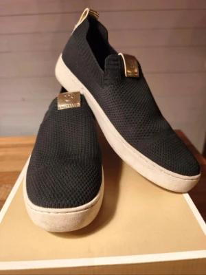 Svarta slip-on sneakers med vit sula - Stilrena svarta slip-on sneakers med stickad ovandel och vit, tjock sula. Skorna har guldfärgade detaljer på plösen och hälen för en extra touch. Perfekta för dig som gillar enkel och modern design. Ovandelen är i mesh-liknande material som andas.