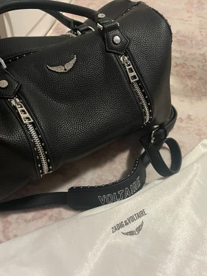 Svart handväska Zadig & Voltaire - Snygg svart handväska i strukturerat läder från Zadig & Voltaire. Väskan har silverfärgade detaljer, dubbla dragkedjor framtill och ett emblem med vingar. Axelremmen är dekorerad med VOLTAIRE i vitt. Perfekt för dig som gillar rockig stil.