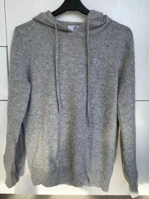 Ljusgrå hoodie med broderad logga - Mysig ljusgrå hoodie med huva och dragsnören. Tillverkad i mjukt stickat material och har en liten vit broderad logga på bröstet. Långa ärmar och ribbade muddar vid ärmslut och nederkant. Perfekt för en chill och avslappnad stil. Alltid kalltvättad och köpt ungefär typ 6 månader sen med väldigt fin skick