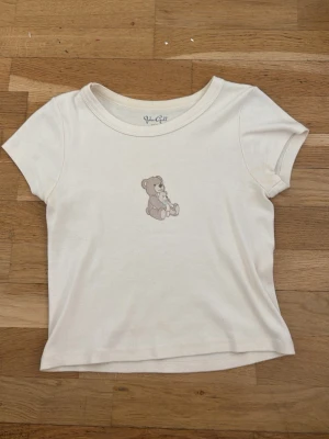 Vit t-shirt med nalletryck - Söt vit t-shirt med korta ärmar och ett gulligt tryck av två nallar på bröstet. T-shirten har en klassisk rund halsringning och är gjord i mjuk bomull som känns skön mot huden. Perfekt för en avslappnad och lekfull stil.