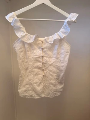 Vit broderad blus med volang - Söt vit blus i bomull med broderade blommor och volang runt axlarna. Ärmlös modell med knappar framtill och romantisk känsla. Perfekt för sommarens alla tillfällen. Dessvärre står det ingen storlek pga att den är begagnad.. men skulle uppskatta den som en XS,32… hör av er ifall ni blir osäkra så skickar jag mått på toppen.💗