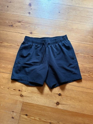Hollister badshorts  - Säljer ett par svarta shorts med elastisk midja och snörning. Shortsen har två sidofickor och är tillverkade i ett lätt, strukturerat material som passar perfekt för varma dagar.