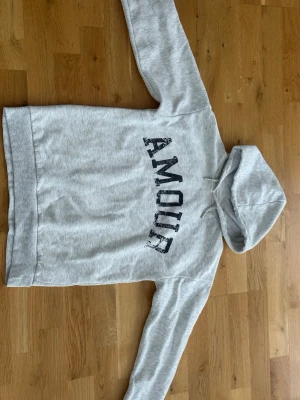 Hoddie  - Ljusgrå hoodie med svart texten 'AMOUR' på framsidan och en stjärna på ryggen. Hoodien har huva med dragsko och är tillverkad i mjukt sweatshirtmaterial. Perfekt för en avslappnad och trendig look.