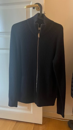 Mörkblå Stickad Helzip från Jack and Jones - Säljer en svart ribbstickad ziptröja med hög krage och långa ärmar. Tröjan har dragkedja framtill och två fickor. Perfekt för lager på lager och enkel att matcha med olika outfits.