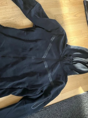 Nike tech  - Säljer en svart Nike tech hoodie med hel dragkedja framtill och huva. Hoodien har diskreta detaljer i form av två diagonala linjer på bröstet och en ficka med dragkedja på ärmen. Perfekt för en avslappnad och sportig stil. Använd fåtal gngr.