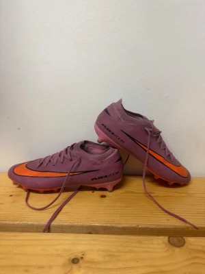 Nike fotbollsskor - Säljer ett par lila Nike fotbollsskor med orange sula och detaljer. Skorna har snörning, låg profil och är tillverkade i syntetmaterial för bra grepp på planen. Swoosh-logga i svart och orange på sidorna samt texten 'AIR ZOOM'. Perfekta för dig som vill sticka ut på fotbollsplanen.