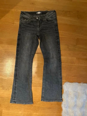 Grå bootcut jeans med ficklock - Säljer ett par grå jeans med bootcut-modell och ficklock bak med knappdetalj. Jeansen har en tvättad look och raka ben som slutar i en lätt utsvängning. Klassisk femficksdesign och sydda detaljer. Perfekta för dig som gillar en avslappnad men ändå trendig stil.