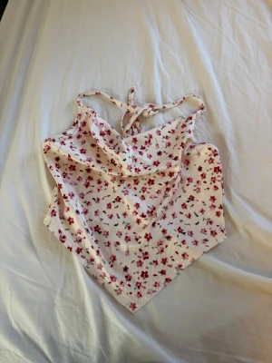Blommig topp med knyt - Supersöt topp med knyt i nacken. Vit botten med små röda blommor och ribbad struktur. Perfekt till sommaren och ger en fräsch vibe till din outfit. Sitter tight och är ärmlös för en luftig känsla.
