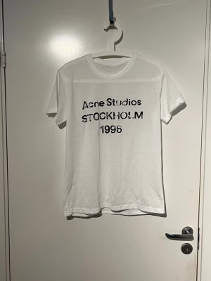 Vit Acne Studios t-shirt - Säljer en vit t-shirt från Acne Studios med svart tryck på bröstet där det står 'Acne Studios STOCKHOLM 1996'. Klassisk passform och rund halsringning. Perfekt till jeans eller shorts för en clean och stilren look.