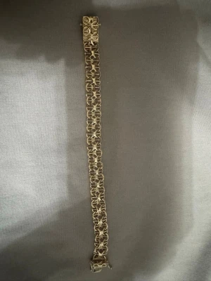 Guldigt X-länk armband  - Snyggt armband i guldfärgad metall med ett unikt, flätat kedjemönster. Armbandet har en bred design och ett dekorativt spänne som ger en exklusiv känsla. Perfekt för dig som vill ha ett statement-smycke till din outfit.