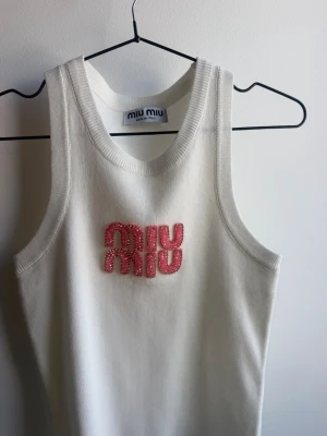 Vit stickad linnetopp från Miu Miu - Supersnygg vit linnetopp från Miu Miu med rosa pärlbroderat logotyp framtill. Toppen är stickad och har breda axelband och rundad halsringning. Perfekt för dig som vill ha en trendig och exklusiv look.