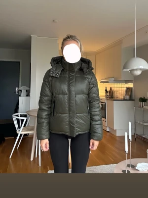 Mörkgrön pufferjacka från KHUJO - Snygg mörkgrön pufferjacka med avtagbar huva och hög krage. Jackan har dragkedja framtill, två sidofickor med dragkedja och quiltad design. Perfekt för kalla dagar och ger en trendig look. Passar till det mesta och är riktigt varm. Väldigt bra skick! Knappt använd! Nypris runt 2000!