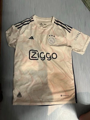 Ajax Amsterdam bortatröja Adidas - Snygg Ajax Amsterdam bortatröja från Adidas i ljusbeige med marinblå detaljer och Ziggo-logga på bröstet. Tröjan har korta ärmar, rund hals och tre stjärnor ovanför klubbmärket. Tillverkad i lätt och ventilerande material, perfekt för fotbollsträning.