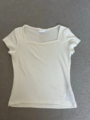 Beige kortärmad topp med fyrkantig ringning - Säljer en enkel och stilren beige topp med korta ärmar och fyrkantig ringning. Toppen har en slim passform och är gjord i mjuk bomull, perfekt till sommaren. Passar till både jeans och kjol för en clean look.